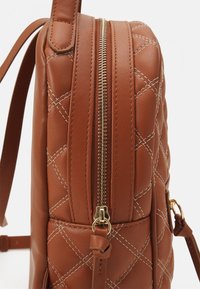 Mochila de cuero marrón acolchada con cremalleras y herrajes dorados, con costuras diagonales y una correa ajustable para un uso variado.