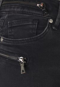 Jeans de mezclilla negra con dos bolsillos frontales y un bolsillo para monedas con cremallera y herrajes plateados. Textura suave y diseño ajustado.