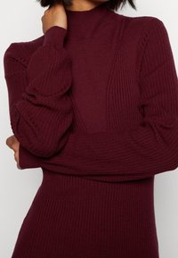 Pull en maille côtelée bordeaux avec un col montant, des manches longues et des détails texturés aux épaules et sur les côtés, offrant une coupe ajustée.