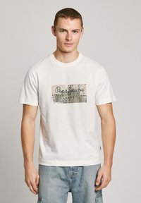Camiseta blanca de algodón con un estampado gráfico rectangular que presenta el texto "Pepe Jeans" en negro, sobre un fondo beige texturizado.