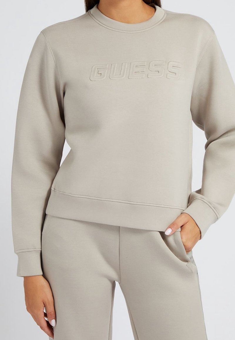 Beige collegepaita, jossa on pyöreä kaula-aukko, ribbireunat ja kohokuvioitu logo edessä. Materiaali näyttää pehmeältä ja sileäpintaiselta.