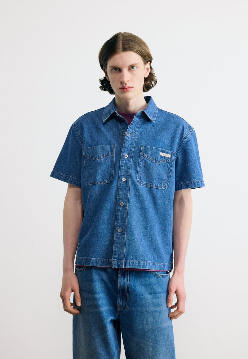 Νέος άνδρας που φοράει κοντομάνικο τζιν πουκάμισο με δύο τσέπες στο στήθος και τζιν παντελόνι σε απόχρωση που ταιριάζει, στέκεται μπροστά σε λευκό, απλό φόντο.