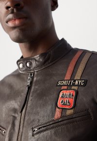 Barna bőrkabát, snap gallérral, elülső cipzárral és két cipzáras zsebel. Patches "SCHOTT-NYC" és "FUEL OIL" részletekkel.
