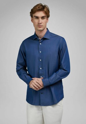 Young man wearing a long-sleeve dark blue button-up shirt and white pants, standing with hands clasped in front against a neutral background.