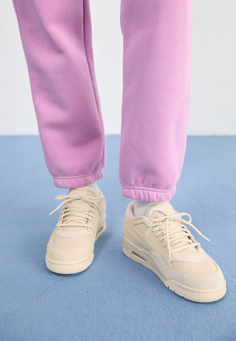 Beżowe sneakersy z teksturowaną cholewką, wyściółką na języku i sznurowanym zapięciem, w zestawie z dopasowanymi różowymi dresami z prążkowanymi mankietami.