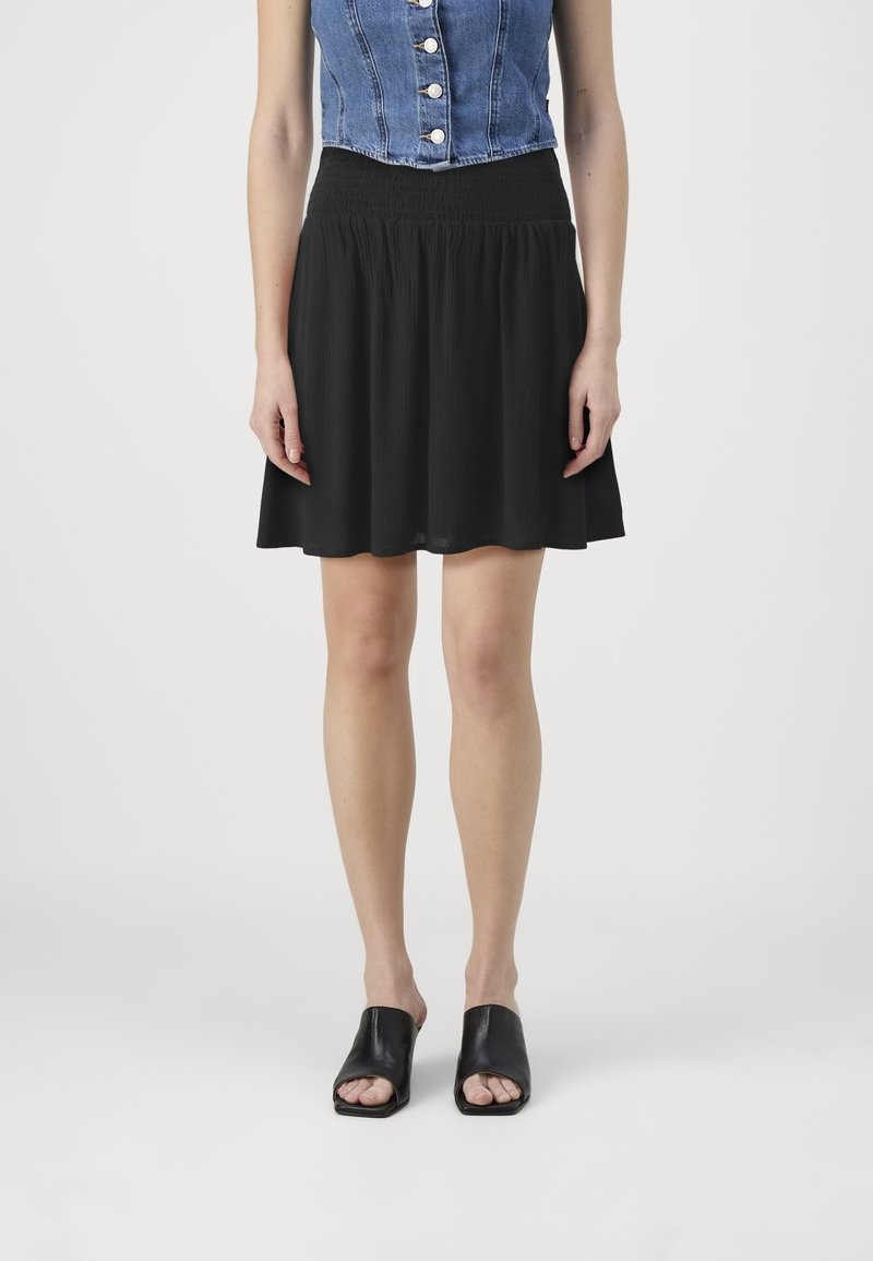 Jupe plissée noire à taille smockée, associée à un haut court en denim. Le mannequin porte des mules noires à bout ouvert. Fond blanc.