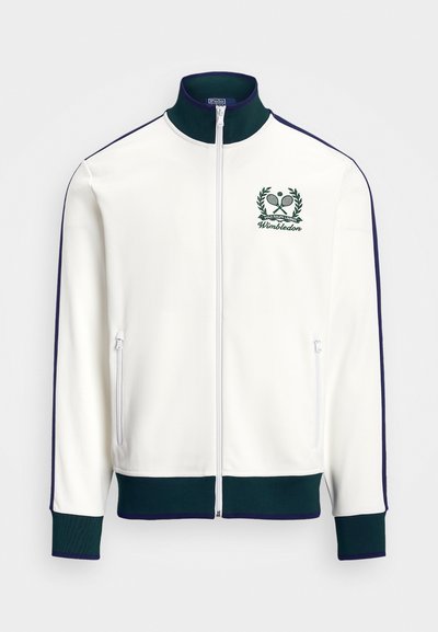 Balts rāvējslēdzēja žaketē ar zaļiem akcentiem, augstu apkakli un Vimbldonas logo ar tenisa motīvu. Iekļautas divas sānu rāvējslēdzēja kabatas.