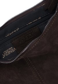 Brązowa zamszowa torba z zamkiem błyskawicznym na górze i kontrastowymi przeszyciami, z wewnętrzną podszewką z tkaniny oraz widoczną markową metką.