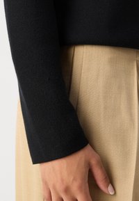 Main posée à côté d'un pantalon plissé beige, portant un haut à manches longues noir texturé.