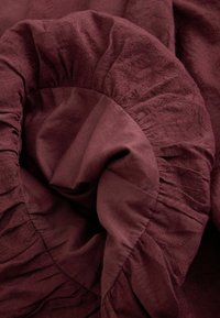 Tela de sábana ajustable de color rojo oscuro estirada y arrugada con banda elástica visible y sutiles patrones de textura floral.