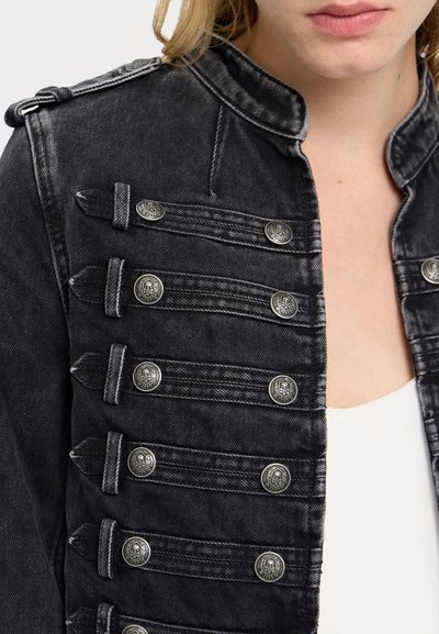 Juodos džinsinės striukės arti su dekoratyviniais sidabriniais mygtukais ir dirželiais, dėvima virš balto marškinėlių ant žmogaus su šviesiais plaukais.