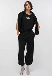 Conjunto negro que consta de una camiseta holgada con logo y pantalones de corte relajado, combinados con sandalias plateadas de tacón, sobre un fondo neutro.