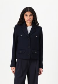 Veste cropped bleu marine avec un col structuré, des boutons croisés et deux poches avant. Texture lisse avec des accents métalliques.