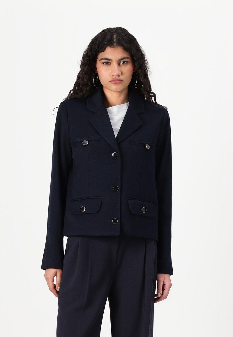 Veste cropped bleu marine avec un col structuré, des boutons croisés et deux poches avant. Texture lisse avec des accents métalliques.