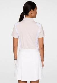 Kobieta z ciemnym kucykiem, ubrana w beżową i białą koszulkę polo oraz białą plisowaną spódnicę, stojąca tyłem na jednolitym tle.