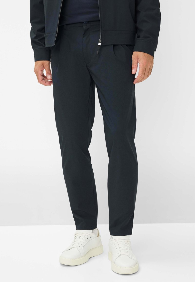 Zwarte, op maat gemaakte broek met een vlakke voorkant, zijzakken en een slim fit, gecombineerd met witte sneakers. Glad materiaal en minimale details.