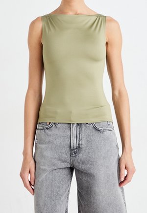 Persona que lleva una camiseta sin mangas de color verde oliva con escote de barco y vaqueros gris claro de talle alto sobre un fondo liso.