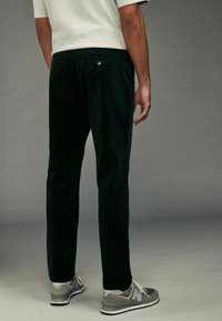Pantaloni din corduroy verde închis cu croi drept, având buzunare pe spate și butoane din metal, asortați cu adidași gri.