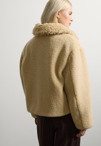 Giacca in pile beige e soffice con superficie testurizzata, ampio colletto e vestibilità rilassata. Le maniche e l'orlo sono leggermente arrotondati.