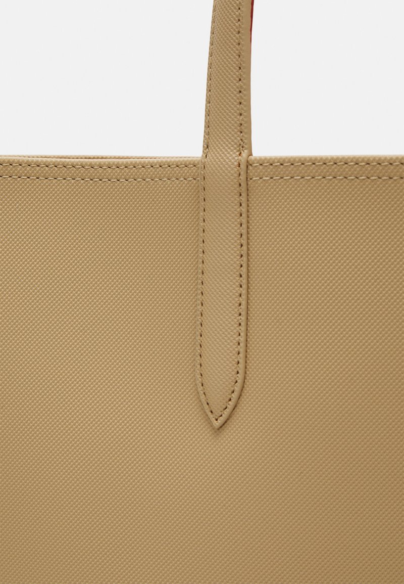 Bolso tote beige con material texturizado y resistente, que presenta una asa delgada y detalles de costura visibles. Diseño simple y rectangular.