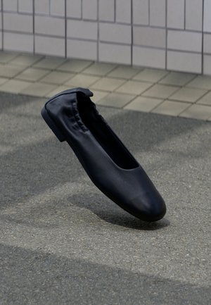 Ballerine en cuir noir avec un dos élastiqué reposant sur un pavement extérieur texturé avec un mur carrelé en arrière-plan.
