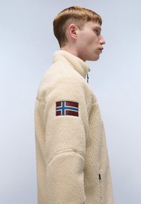 Cream fleece jacket with high collar and zipper, featuring a patch with a blue and red flag on the left sleeve. Textured fabric visible.