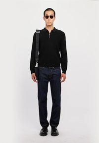 Polo shirt nero a maniche lunghe con colletto con cerniera, abbinato a jeans in denim scuro e scarpe nere, completato con occhiali da sole rotondi e borsa a tracolla.