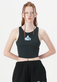 Débardeur noir court avec une texture côtelée, arborant un logo "OBEY" blanc sur le devant. Le modèle le porte avec un pantalon noir ample.