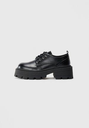 Chaussure en cuir noire à lacets avec semelle épaisse à crampons et petit talon bloc, vue de côté sur un fond clair uni.