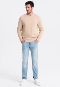 Sudadera beige de cuello redondo en tejido suave; combinada con jeans azul claro; llevada con zapatillas blancas; corte relajado, diseño simple.