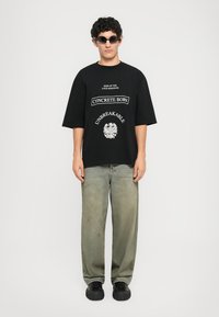 Svart oversized t-skjorte med hvit tekst og grafikk, kombineres med løse, falmede grønne jeans og svarte joggesko. Modellen bærer mørke solbriller.