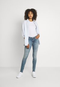 Camisa de botones azul claro sobre una camiseta blanca, combinada con jeans skinny azules desgastados y zapatillas blancas. Cabello rizado y manos en los bolsillos.