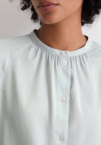 Jasnoniebieska koszula z guzikiem z okrągłym dekoltem, zebrany materiał poniżej dekoltu i białe guziki z przodu. Miękka, gładka tekstura.