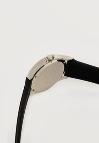 Ανδρικό ρολόι χειρός από ανοξείδωτο ατσάλι με μαύρο λουράκι από σιλικόνη με υφή, εμφανιζόμενο από την πίσω πλευρά, τονίζοντας το περίβλημα του ρολογιού και το κουμπί ρύθμισης ώρας.