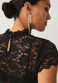 Sort blonder top med høj halsudskæring med flæsekant, korte ærmer og knaplukning bagpå; kombineret med en lang krystallignende ørering.