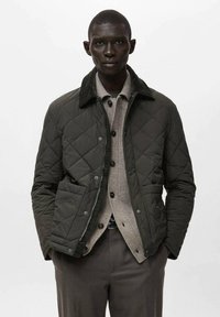 Giacca trapuntata verde scuro con collo in velluto a coste, dotata di bottoni a pressione, due tasche frontali e chiusura con zip, indossata sopra un maglione grigio chiaro.