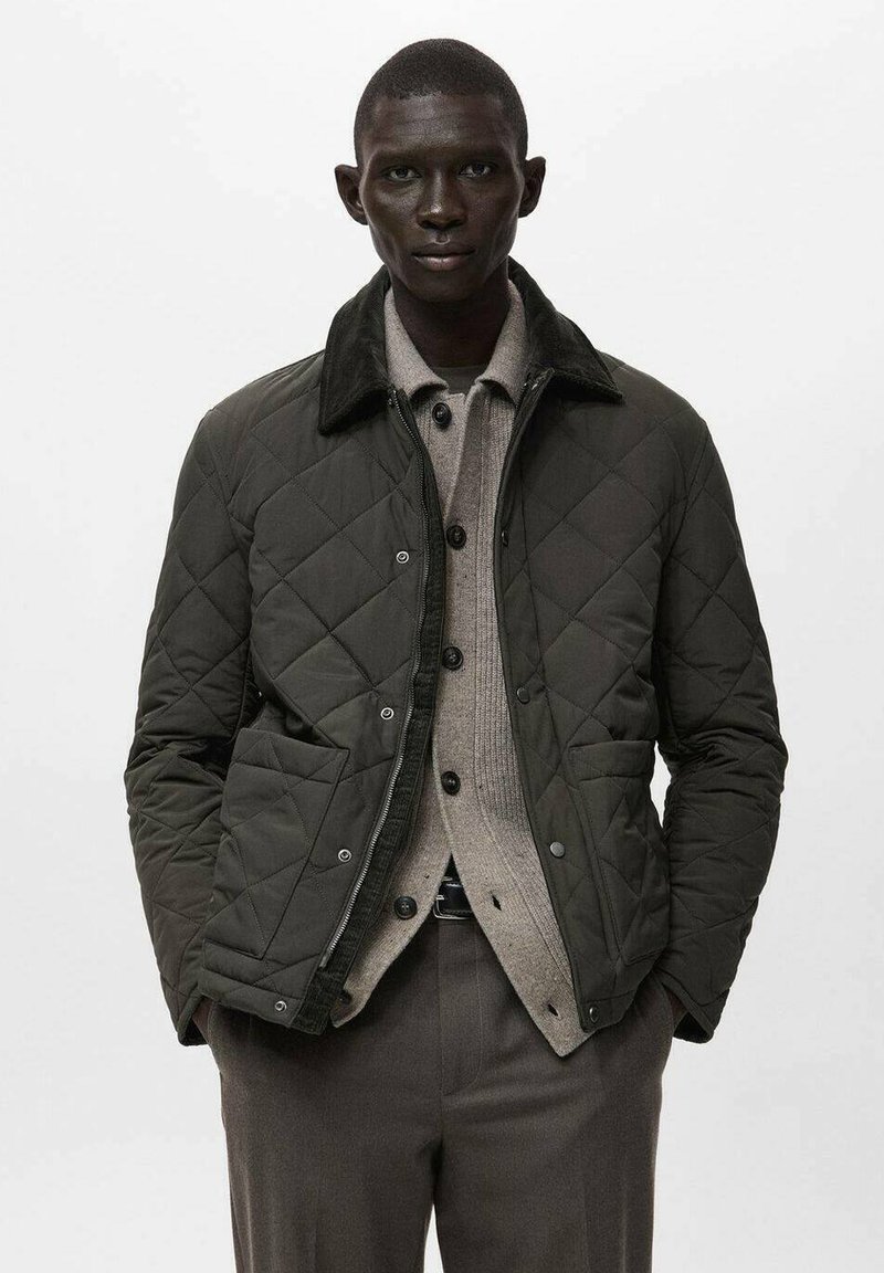 Giacca trapuntata verde scuro con collo in velluto a coste, dotata di bottoni a pressione, due tasche frontali e chiusura con zip, indossata sopra un maglione grigio chiaro.
