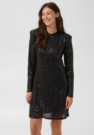 Femme souriante portant une robe noire à manches longues avec des sequins et un col rond sur un fond uni gris clair.