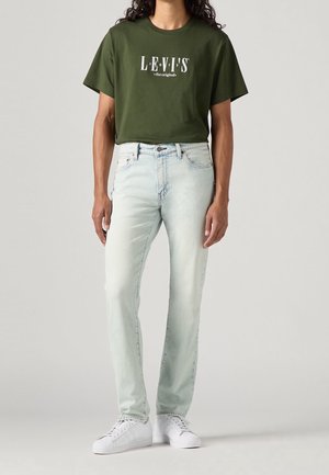 Person trägt ein olivgrünes Levi's T-Shirt, hellblaue Jeans und weiße Turnschuhe und steht vor einem einfarbigen hellgrauen Hintergrund.