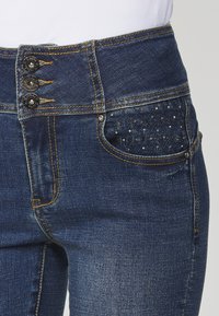 Jean en denim foncé avec une taille haute, une fermeture à triple bouton, des coutures contrastantes et une poche texturée ornée de petits clous.