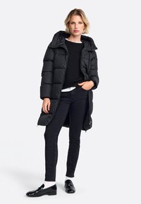 Zwarte pufferjas met een capuchon, met ritssluiting en een horizontaal gequilt design. Gekocht over een zwarte trui en skinny zwarte jeans.