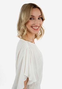 Blusa bianca con maniche corte plissettate e tessuto texturizzato. Collo liscio e vestibilità rilassata. Capelli a caschetto lungo, posa sorridente contro uno sfondo bianco.