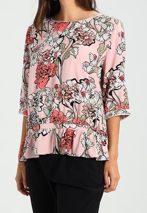 Blouse florale rose, avec des manches courtes, un ourlet peplum et des contours noirs. Le design comprend des fleurs blanches et rouges sur une texture douce.