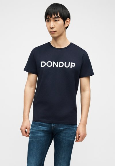 Νεαρός άνδρας που φοράει μπλε μπλουζάκι με λευκό λογότυπο "DONDUP" και μπλε τζιν, στέκεται μπροστά σε μια απλή φωτεινή επιφάνεια.