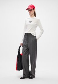 Cămașă albă cu mânecă lungă, cu textul "tommy girl", pantaloni largi cu dungi gri, șapcă roșie și geantă neagră cu accente roșii.