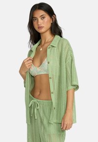 Maglietta verde chiaro a maglia aperta con bottoni, indossata sopra un bikini floreale. L'outfit include pantaloni corti con cordoncino abbinati, con una finitura texturizzata.