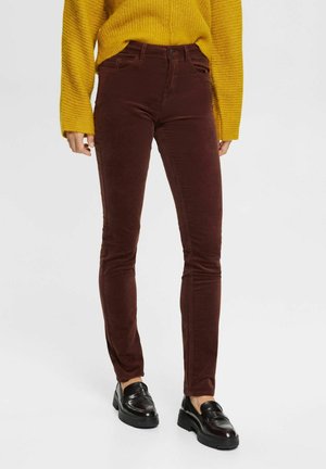Person trägt braune, eng anliegende Cordhosen, schwarze Loafers und einen senfgelben Strickpullover vor einem einfarbigen hellen Hintergrund.