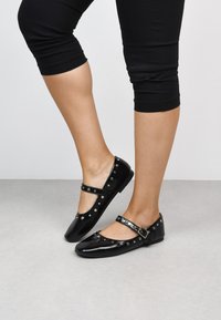 Bailarinas de charol negro con correa ajustable, punta redonda y acentos de tachuelas plateadas a lo largo de los bordes. Acabado suave y tacón bajo.