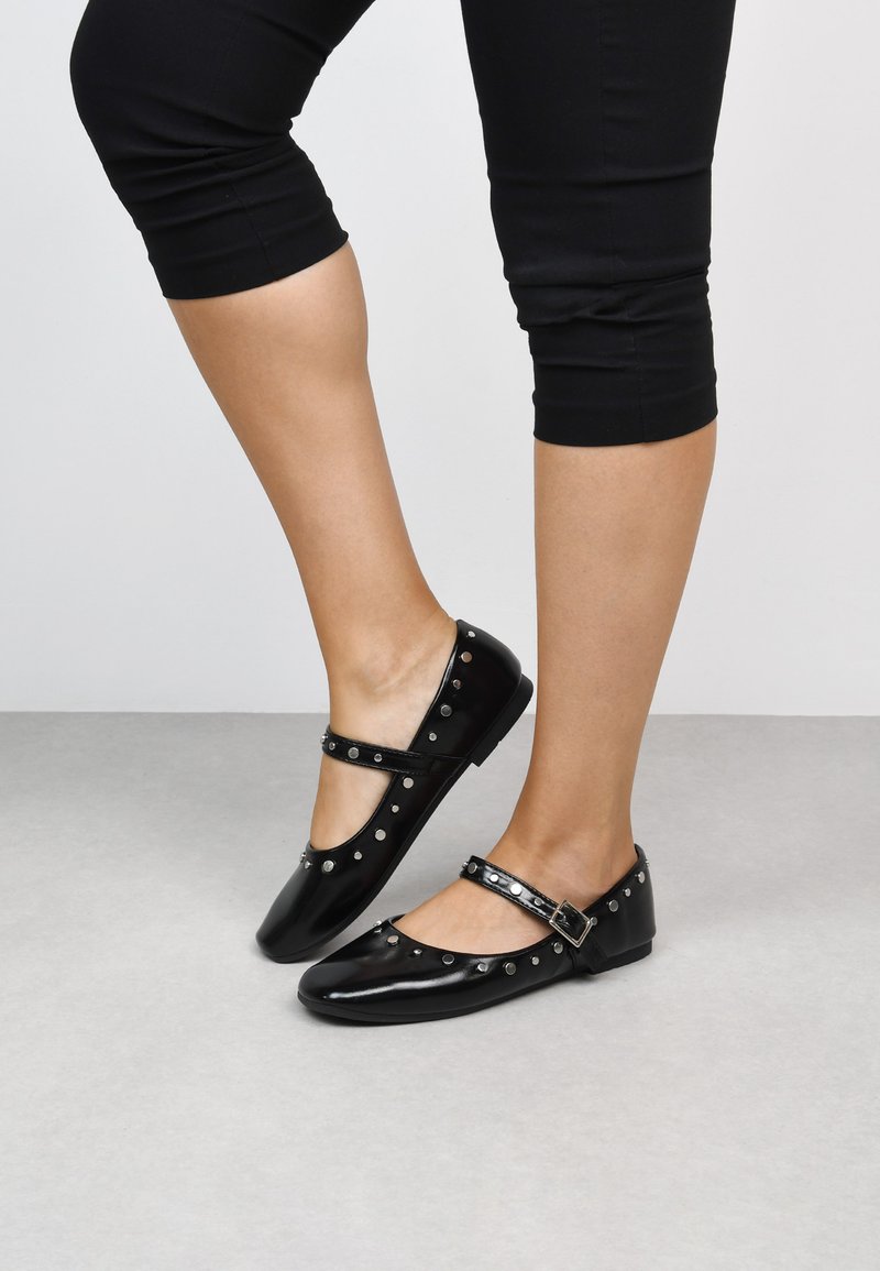 Bailarinas de charol negro con correa ajustable, punta redonda y acentos de tachuelas plateadas a lo largo de los bordes. Acabado suave y tacón bajo.