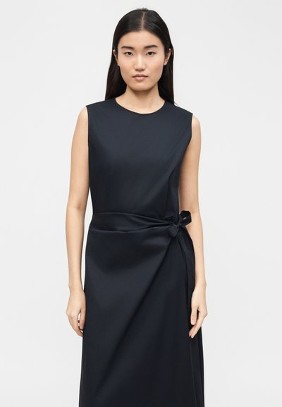 Jeune femme aux longs cheveux noirs portant une robe noire sans manches avec un nœud sur le côté, debout devant un fond gris clair uni.